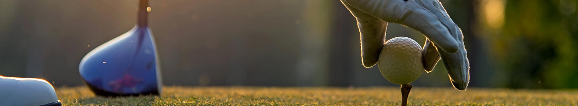 banner image of a golfers hand placing a golf ball onto a tee with focus on the hand and ball while the course and golf club can be seen out of focus in the background.